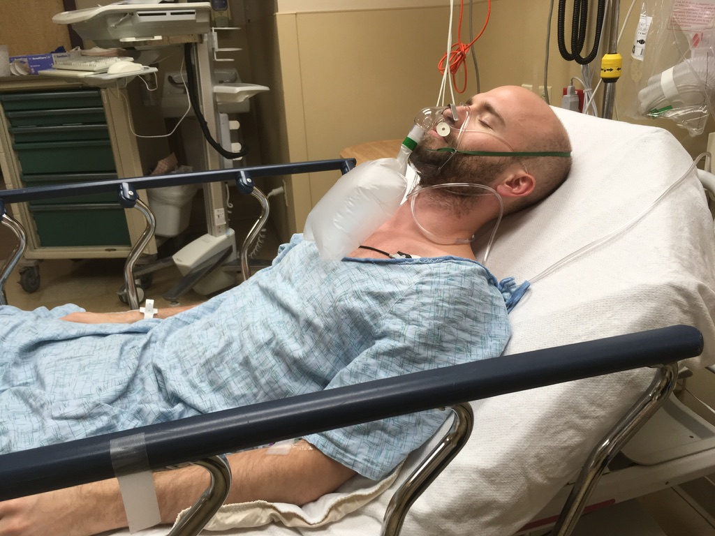 Mikey - Laying in ICU bed, eyes closed, gown worn, oxygen mask on, age 29, medical equipment in background