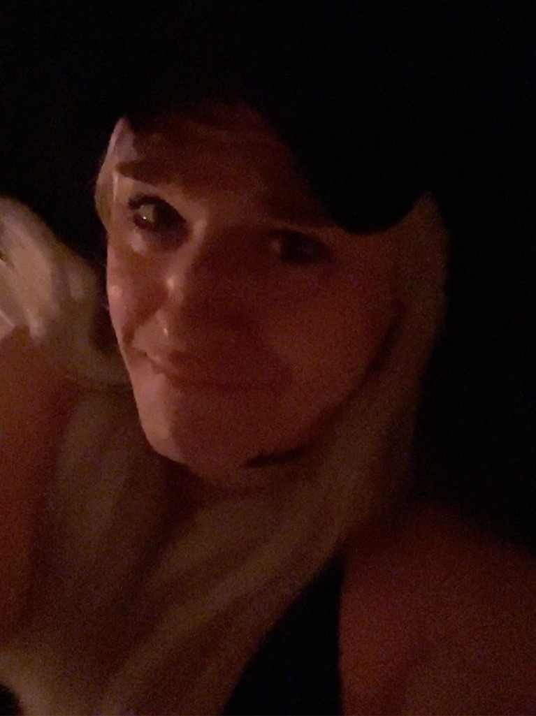 Mikey - Age 34, presenting feminine, laying on soft surface, platinum blonde hair, low light, makeup applied, sorrowful expression, wearing: black ball cap, black sports bra